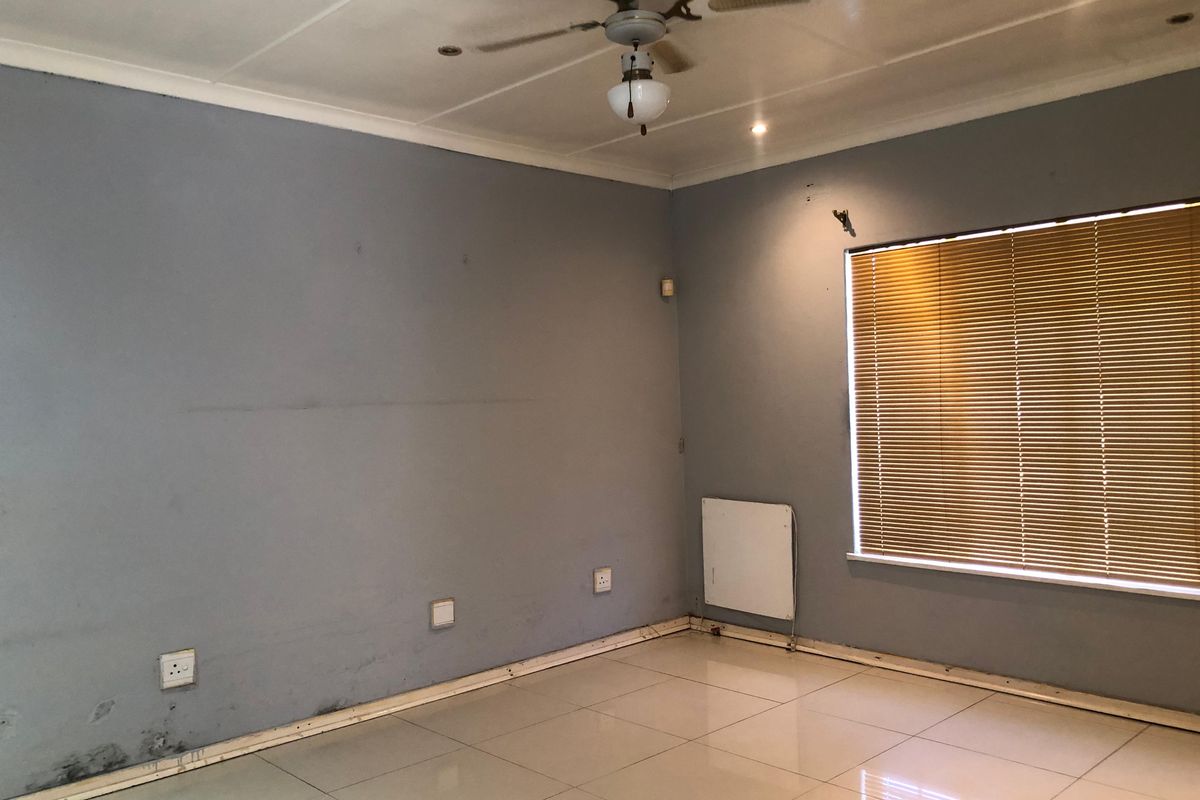 An empty living room featuring tiled floors, a ceiling fan, and venetian blinds, showing signs of wear and potential moisture damage at the wall base.