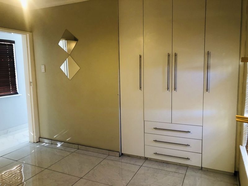 A clean, empty bedroom featuring modern white built-in cupboards, neutral wall tones, and polished floor tiles with natural light from the window.