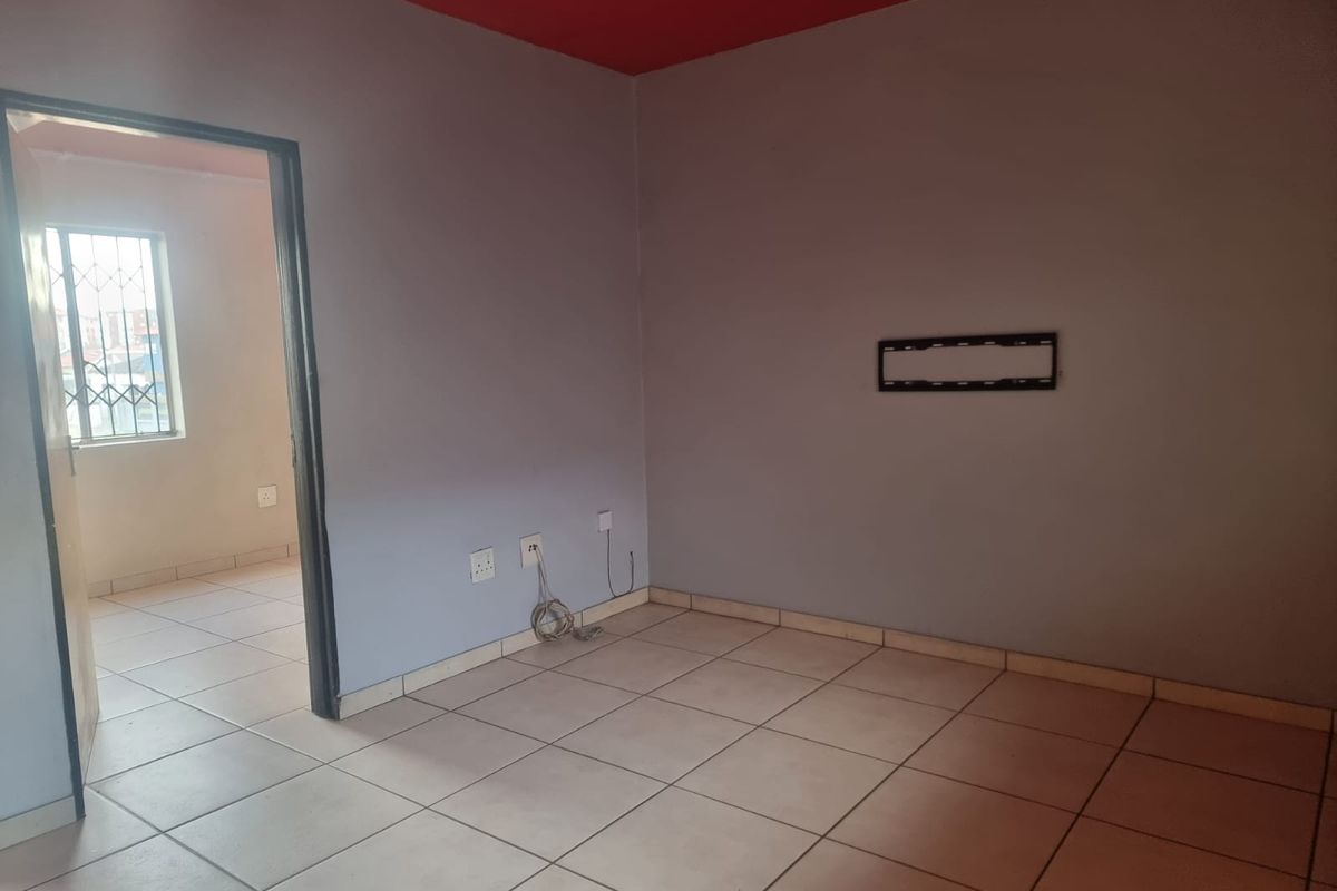 An empty living room featuring tiled flooring, a wall-mounted TV bracket, and a doorway leading to an adjacent room with security-barred windows.