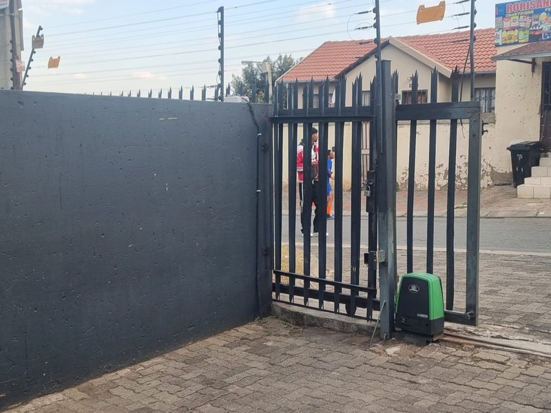 Front entrance featuring a secure sliding gate with an automated motor, perimeter wall with electric fencing, and paved driveway access.