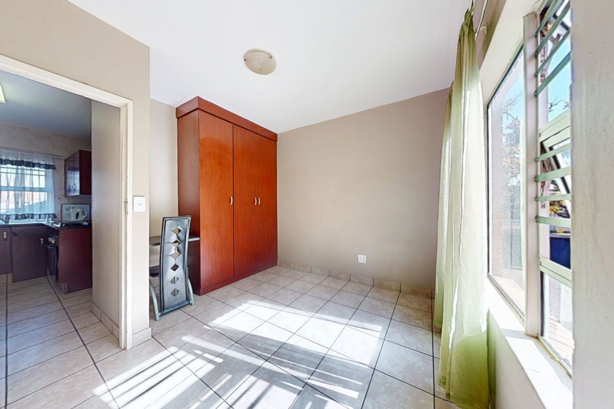A bedroom featuring tiled flooring, a built-in wooden wardrobe, and a window with security bars, with an adjacent view into the kitchen area.