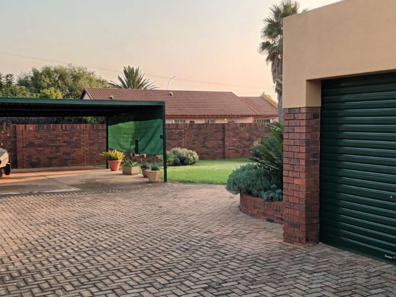 A spacious rear exterior area featuring a paved driveway, a covered carport, and a secure garage with a brick perimeter wall.
