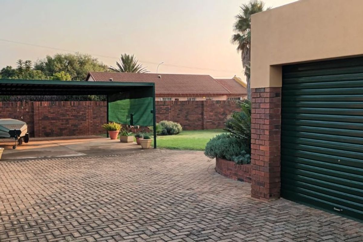 A spacious rear exterior area featuring a paved driveway, a covered carport, and a secure garage with a brick perimeter wall.