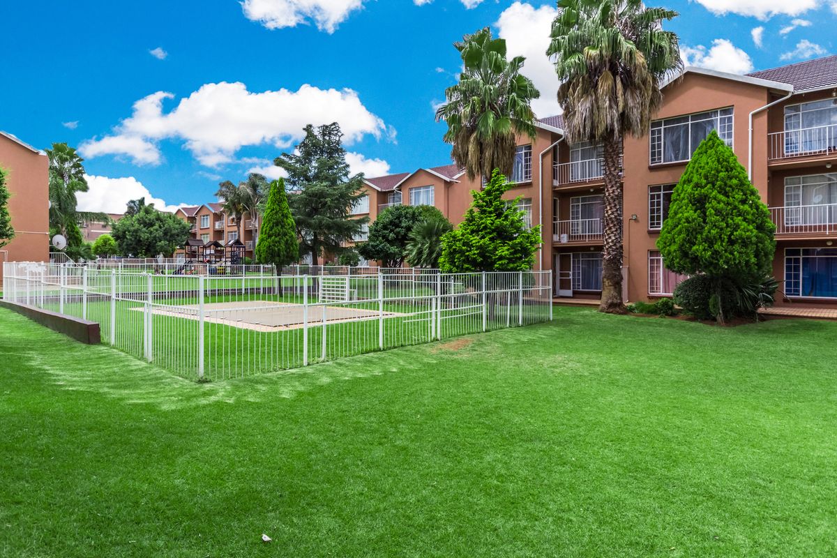 A well-maintained communal garden area featuring a fenced lawn, mature palm trees, and views of the apartment complex facade with private balconies.