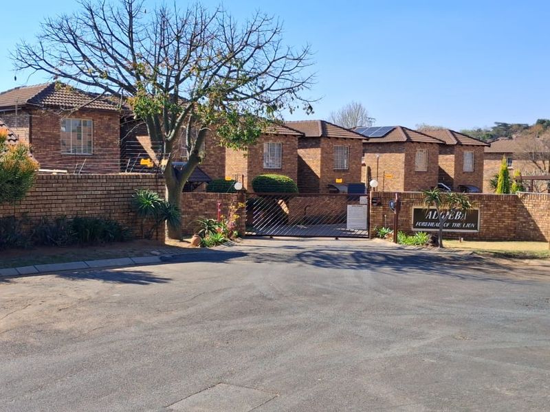 Front exterior view of a brick townhouse complex featuring a secure gated entrance, paved driveway, and solar panel installations on the tiled roofs.