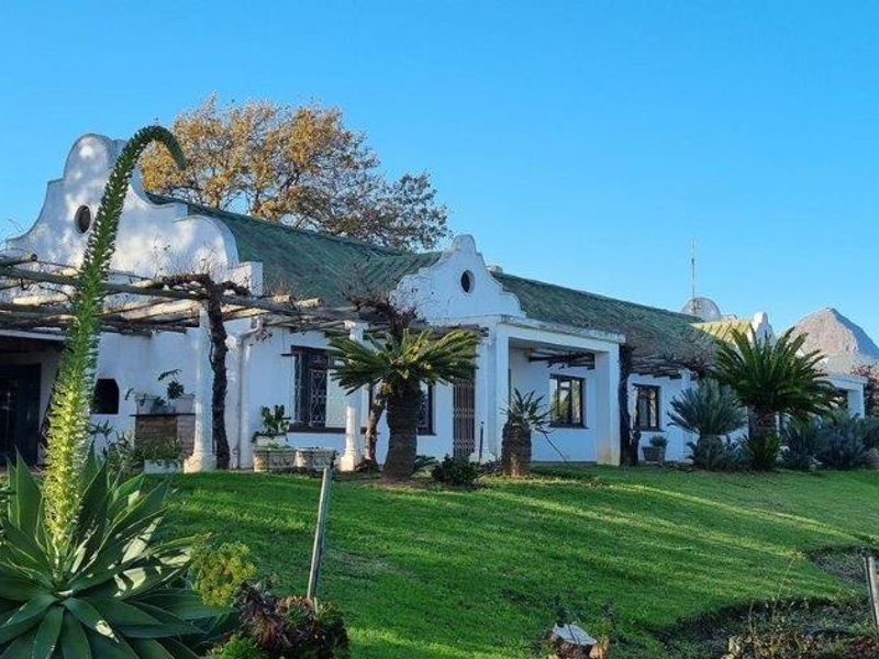 A charming Cape Dutch style farmhouse featuring a covered pergola patio, expansive green lawns, and scenic mountain views in a tranquil rural setting.