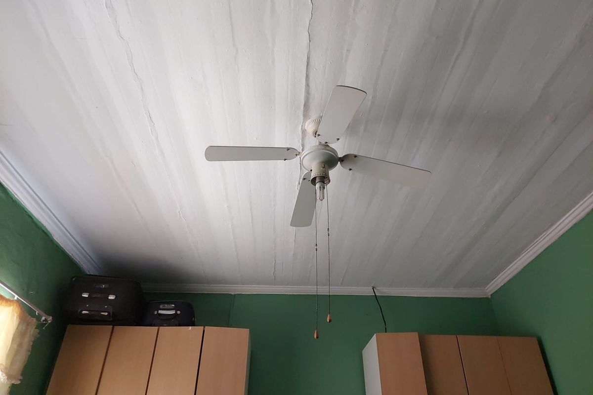 A bedroom interior featuring built-in cupboards and a ceiling fan, showing visible signs of ceiling damage and uneven surface finishes.