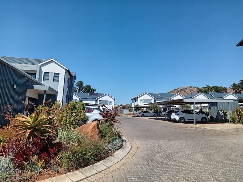 Exterior view of a modern apartment complex featuring paved driveways, covered carport parking, and well-maintained landscaped garden areas under a clear blue sky.