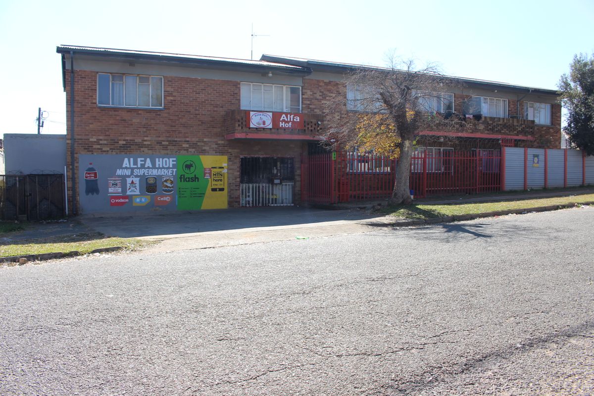 Exterior view of the Alfa Hof apartment building featuring a ground-floor commercial space, brick facade, and perimeter security fencing.