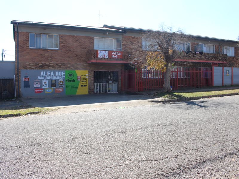 Exterior view of the Alfa Hof apartment building featuring a ground-floor commercial space, brick facade, and perimeter security fencing.