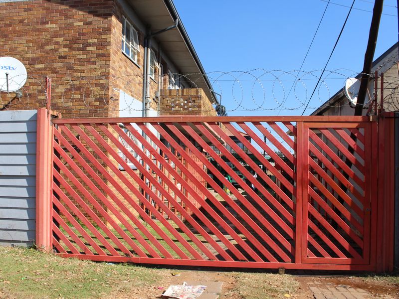 A secure street-facing entrance featuring a red metal sliding gate, brick facade, and perimeter security fencing with razor wire.