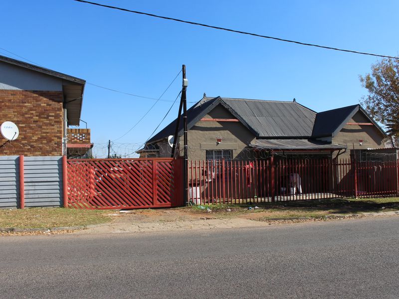 Street view of a residential property featuring a metal perimeter fence, gated entrance, and a single-story building with a pitched roof.