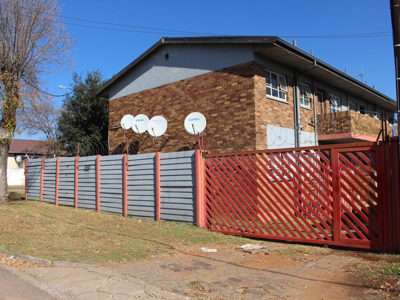 Exterior view of an apartment building featuring a brick facade, metal security gate, and perimeter fencing with mounted satellite dishes.