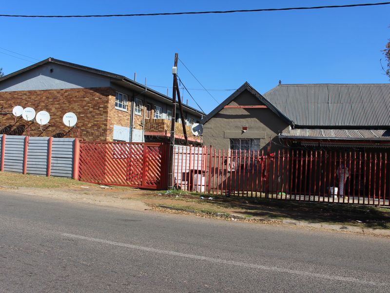 Street view of the property featuring a brick apartment building and adjacent structure, secured by red metal perimeter fencing and gates.