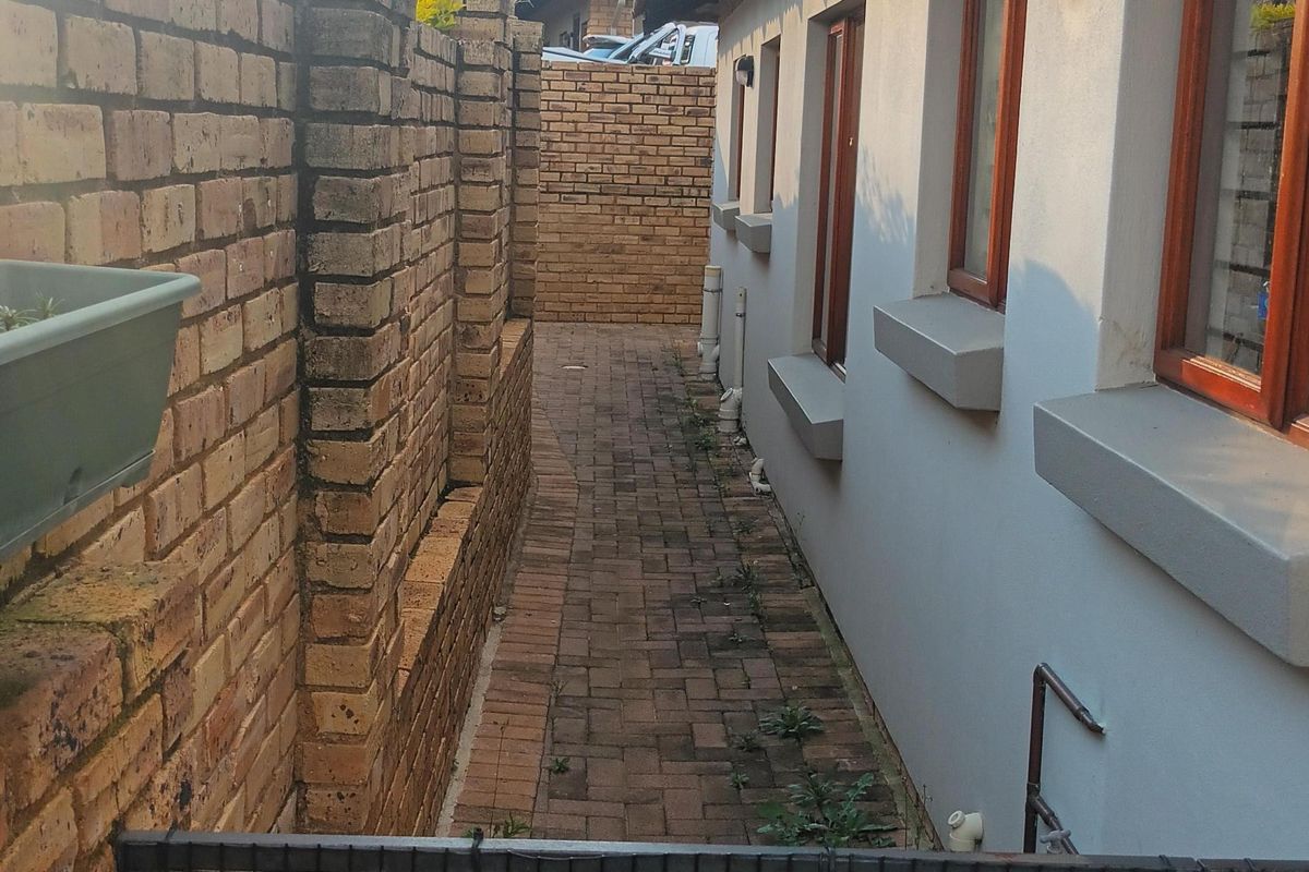 A narrow paved side passage featuring a brick boundary wall, external lighting, and a metal security gate, providing access along the side of the property.