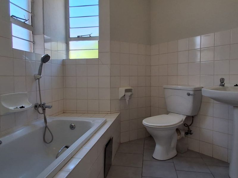 A dated bathroom featuring a built-in bathtub, pedestal basin, and toilet with original wall tiling and louver windows.