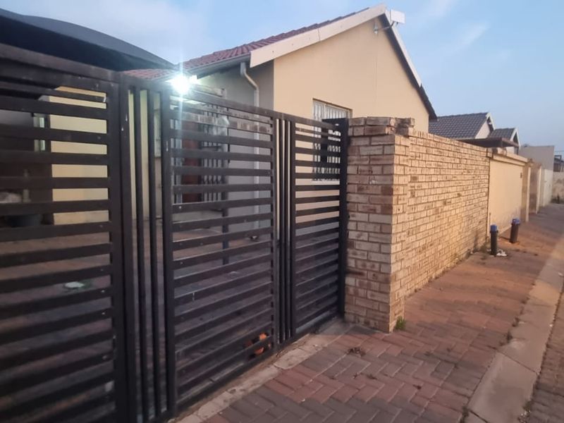 Front exterior view of the property featuring a brick boundary wall, metal driveway gate, and paved entrance area.