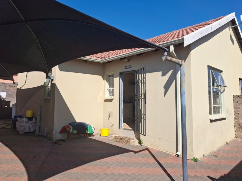 Rear exterior view of the house featuring a paved patio area, shade sail, and secure entrance with a metal security gate.