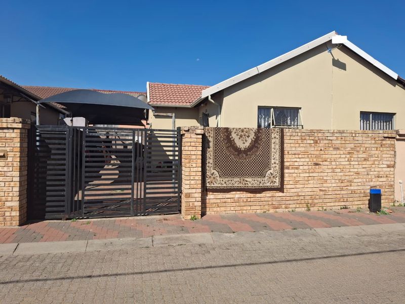 Front exterior view of a single-story house featuring a brick boundary wall, metal driveway gate, and a shade cloth carport.