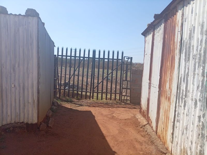 An exterior view showing a metal gate entrance flanked by weathered corrugated iron structures on a dirt path.