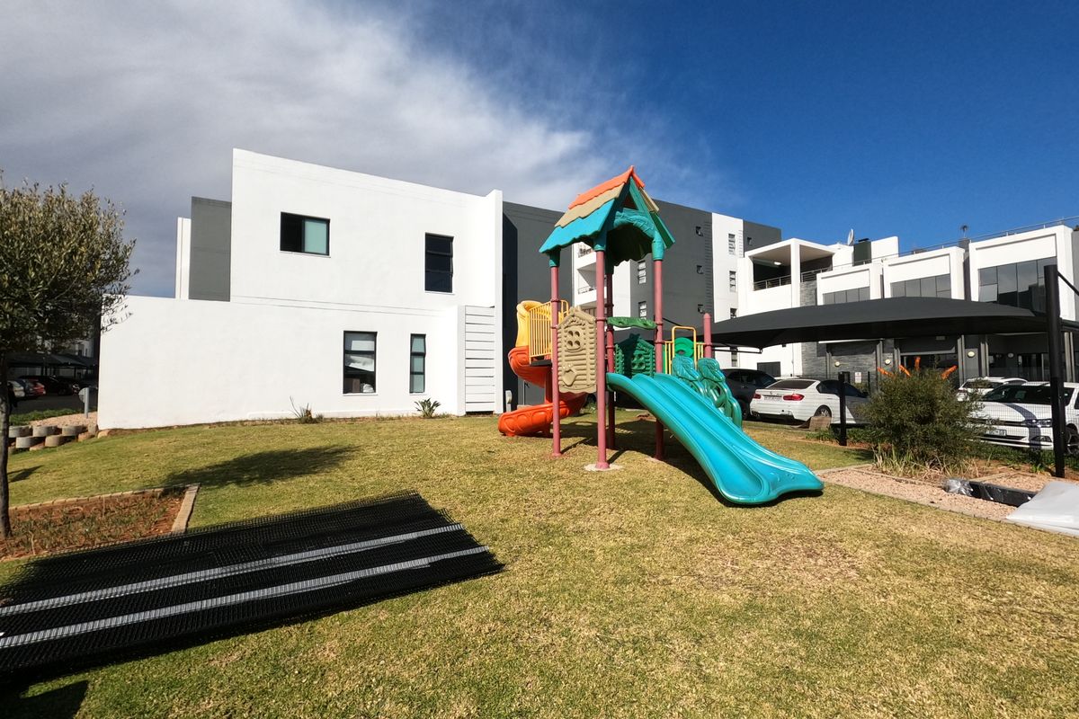 A well-maintained communal garden area featuring a children's playground, paved parking bays with shade sails, and modern apartment building architecture.