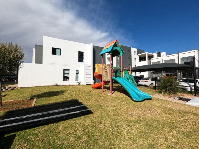 A well-maintained communal garden area featuring a children's playground, paved parking bays with shade sails, and modern apartment building architecture.
