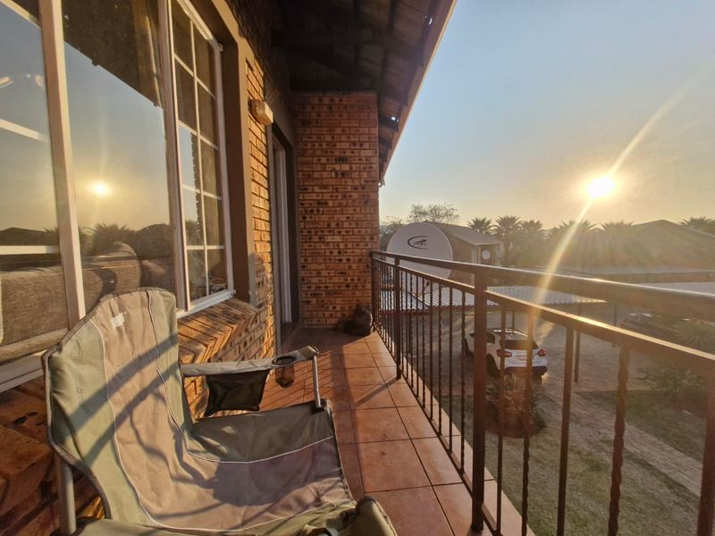 A covered balcony featuring tiled flooring, exposed brick walls, and a metal balustrade, offering an outdoor space with views of the complex.