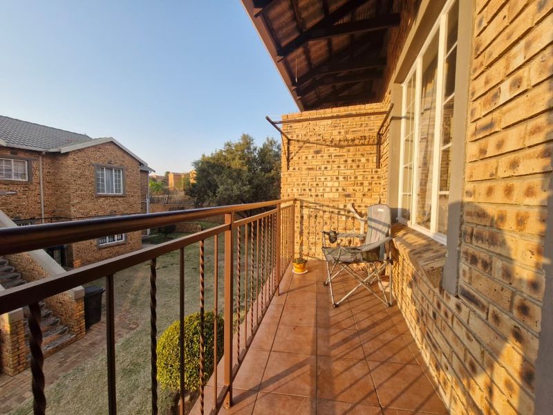 A covered balcony featuring exposed brick walls, tiled flooring, and a metal balustrade, overlooking the apartment complex grounds.