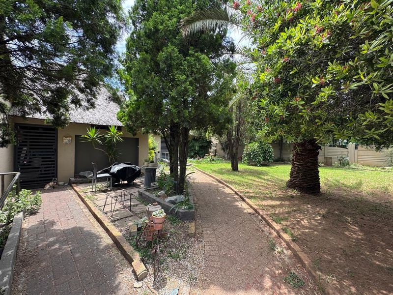 A spacious rear garden featuring mature trees, a paved walkway, and a covered patio area adjacent to the main dwelling.