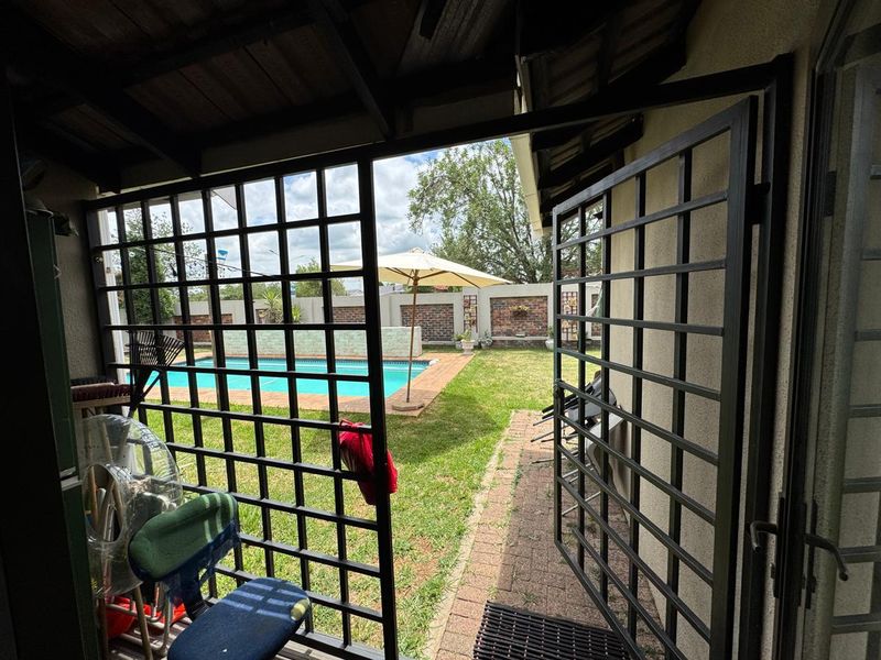 A covered patio area featuring metal security gates, overlooking a private garden with a swimming pool and brick boundary wall.