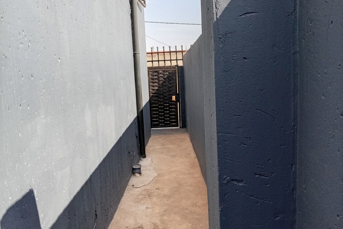 A narrow side passage between two concrete boundary walls, featuring a metal security gate at the end and a concrete pathway.
