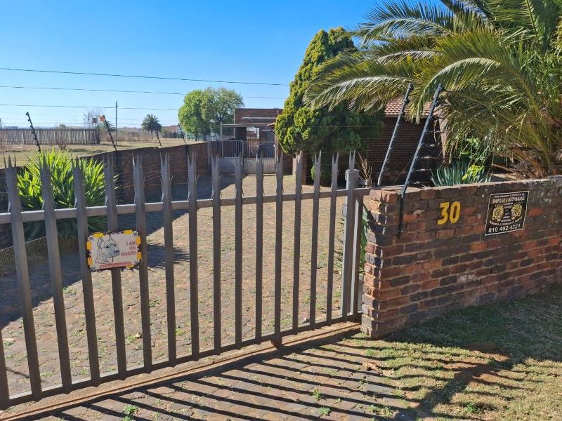 Front view of the property featuring a paved driveway, secure metal gate, brick boundary wall, and electric fencing.