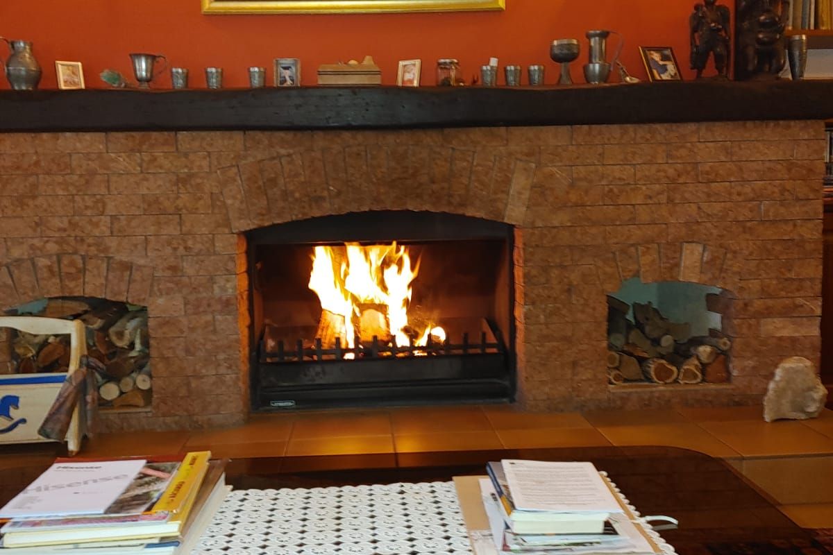 A cozy living room featuring a prominent brick fireplace with a timber mantelpiece, built-in log storage, and warm wall sconce lighting.