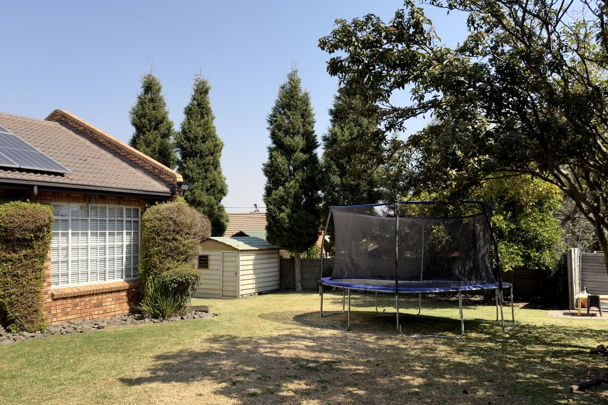 A spacious rear garden featuring a well-maintained lawn, mature trees, a storage shed, and a brick-built house with roof-mounted solar panels.
