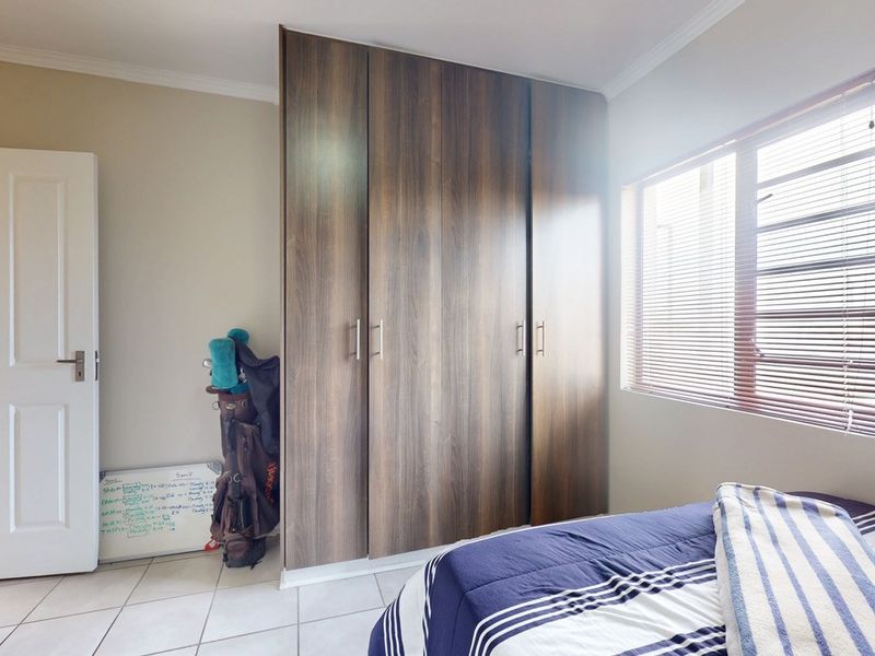 A well-lit bedroom featuring modern wood-finish built-in cupboards, tiled flooring, and a window with fitted blinds.