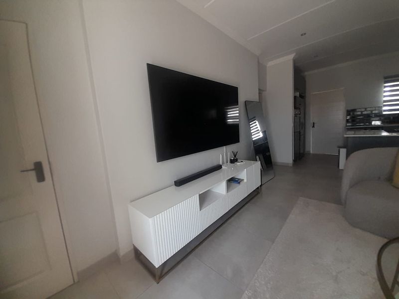 An open-plan living area featuring neutral tiled flooring, recessed ceiling lighting, and a clean, modern aesthetic with a wall-mounted television setup.