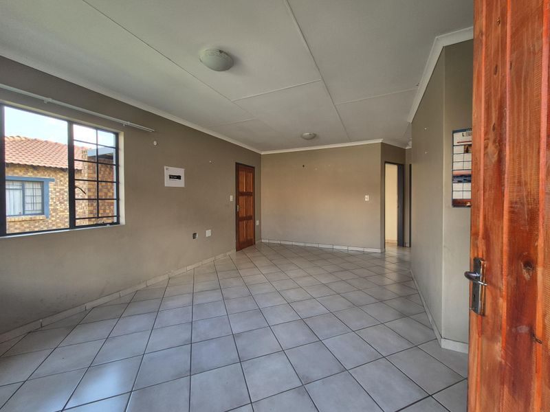 An empty, open-plan living area featuring neutral wall tones, tiled flooring, and a timber entrance door with natural light from the side window.