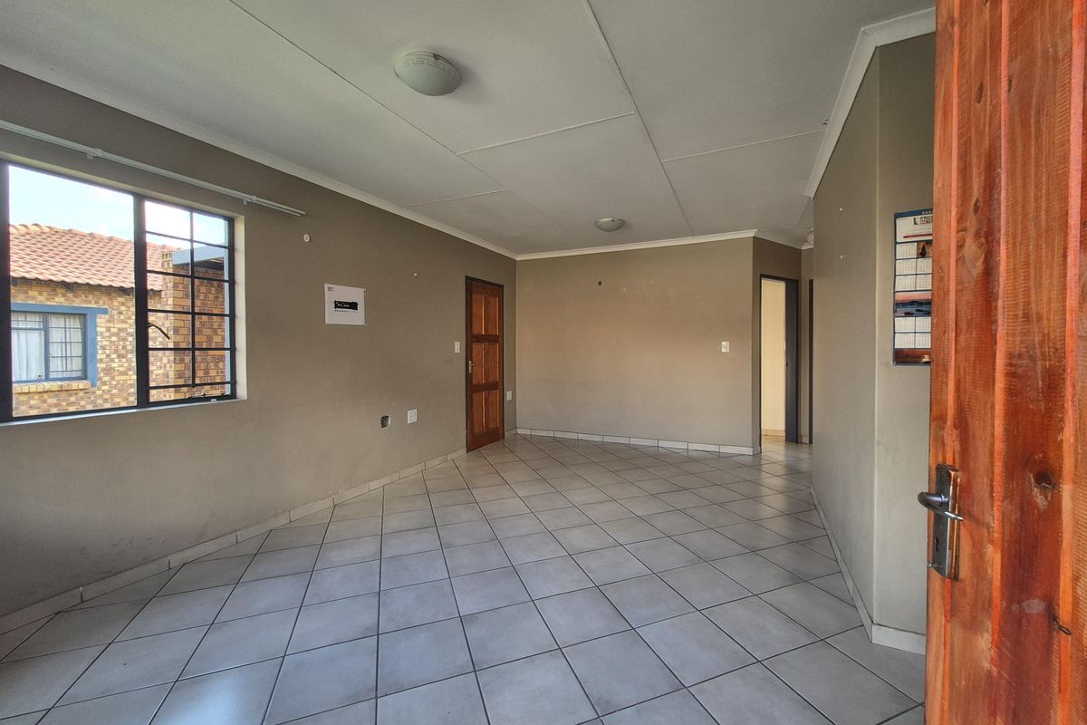 An empty, open-plan living area featuring neutral wall tones, tiled flooring, and a timber entrance door with natural light from the side window.