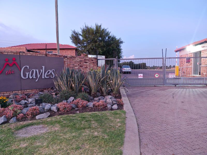 The secure entrance to The Gayles townhouse complex, featuring a gated access point, brick boundary wall, and landscaped garden area.