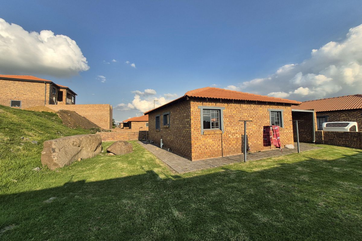 A rear view of a face brick townhouse featuring a tiled roof, paved perimeter walkway, and a well-maintained lawn area.