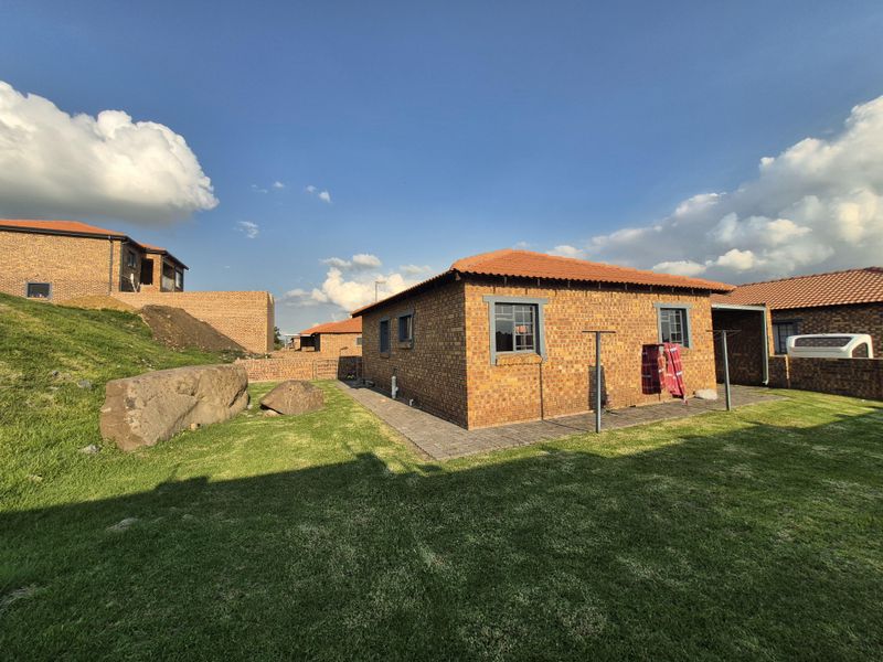 A rear view of a face brick townhouse featuring a tiled roof, paved perimeter walkway, and a well-maintained lawn area.