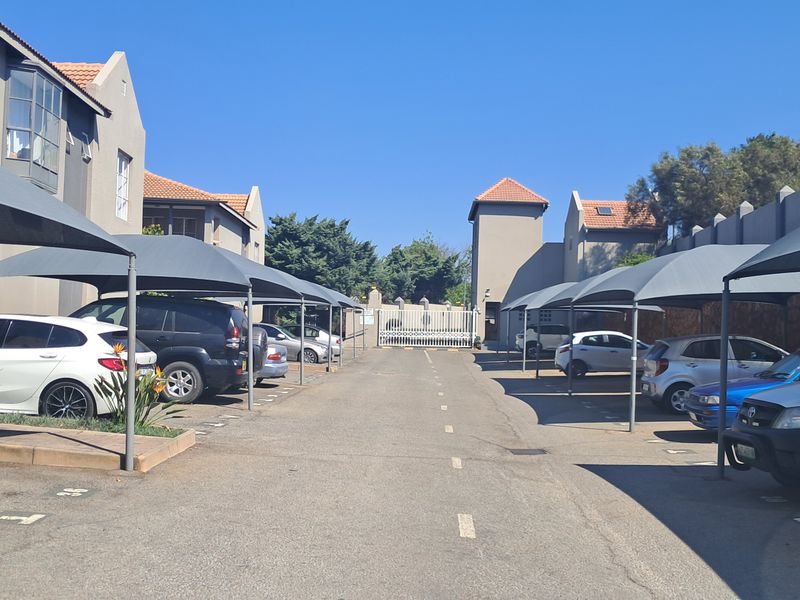 Exterior view of the apartment complex parking area featuring covered shade cloth carports, paved driveway, and secure perimeter fencing.