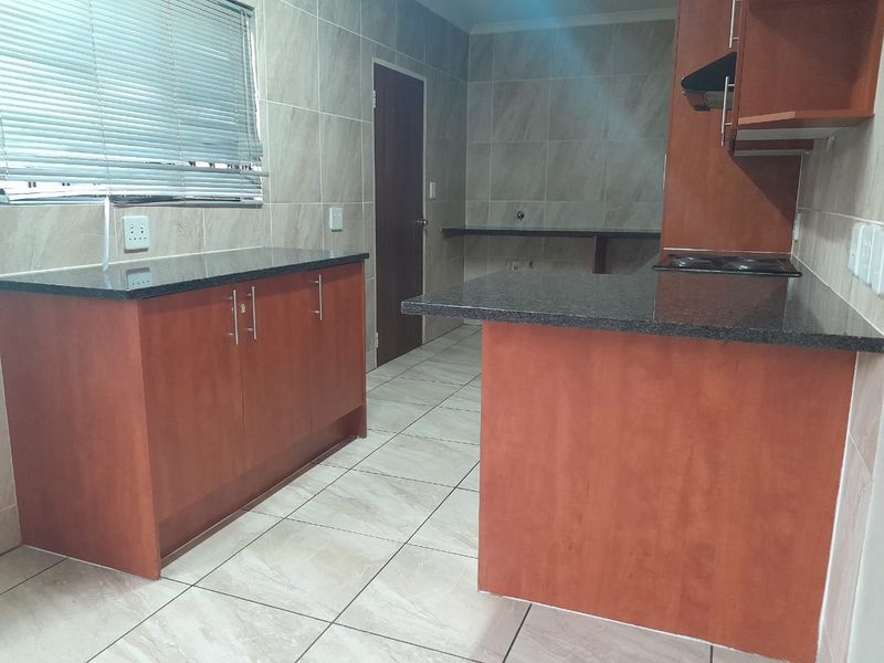 A functional kitchen featuring granite countertops, built-in wooden cabinetry, an electric hob with extractor fan, and neutral tiled finishes throughout.