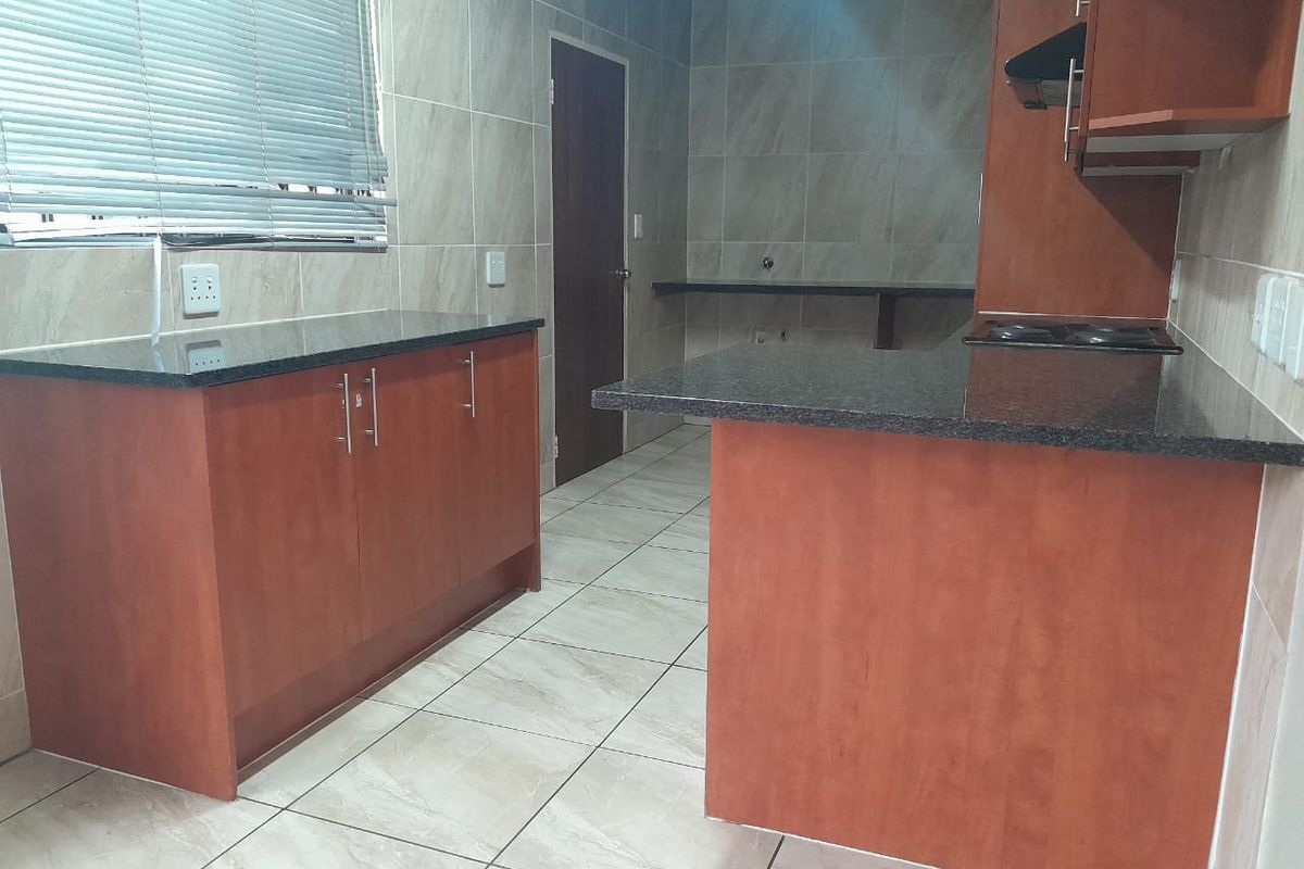 A functional kitchen featuring granite countertops, built-in wooden cabinetry, an electric hob with extractor fan, and neutral tiled finishes throughout.