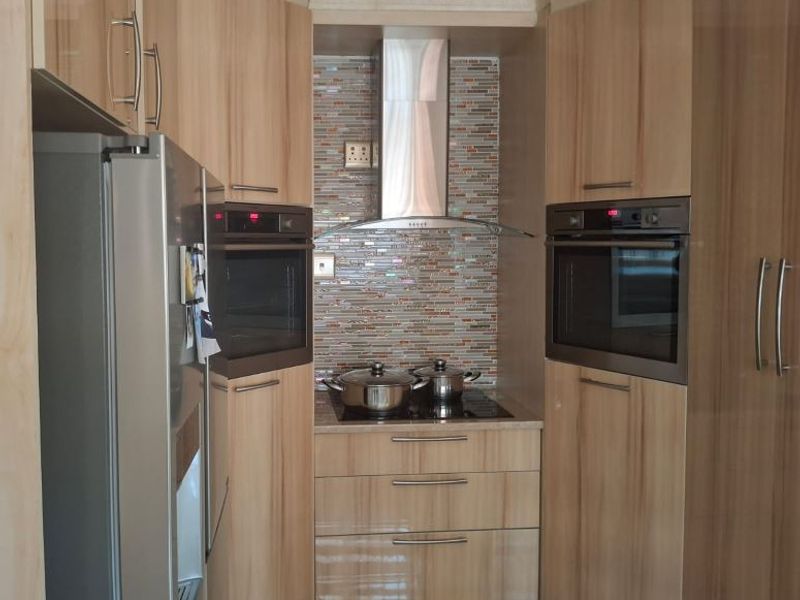 A compact, modern kitchen featuring wood-finish cabinetry, dual built-in ovens, a glass cooktop, and a decorative mosaic tile backsplash.