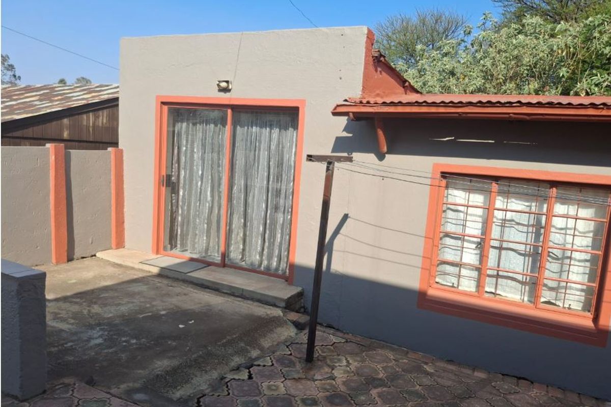 Rear exterior view showing a concrete patio, sliding glass door, and window with security bars, featuring a paved area and corrugated metal roof.