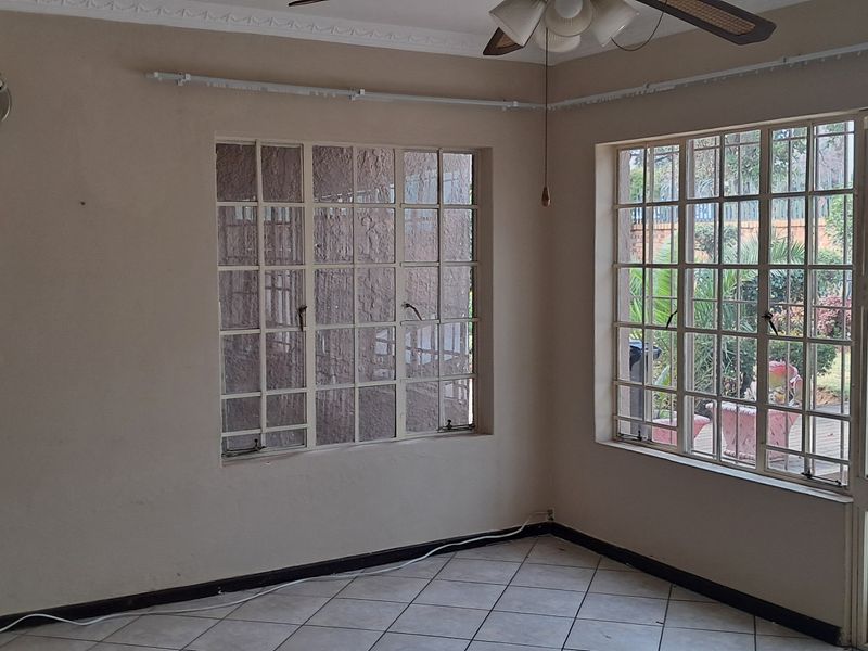 A bright living room featuring tiled flooring, large windows with security bars, and a ceiling fan, offering direct access to the outdoor area.