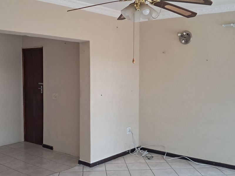 A spacious living area featuring tiled flooring, crown molding, a ceiling fan, and natural light from a window with security bars.