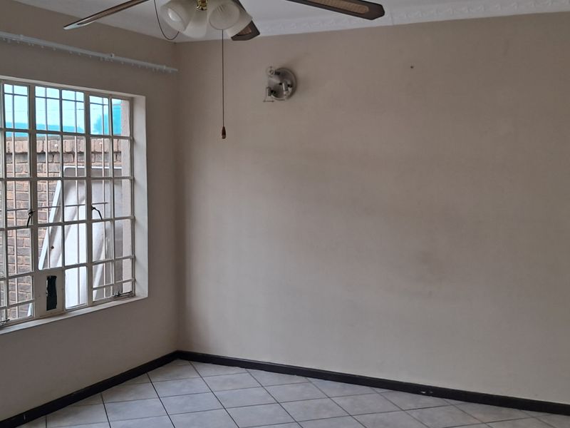 A vacant living room featuring tiled flooring, a ceiling fan, and large windows with security bars overlooking the exterior.