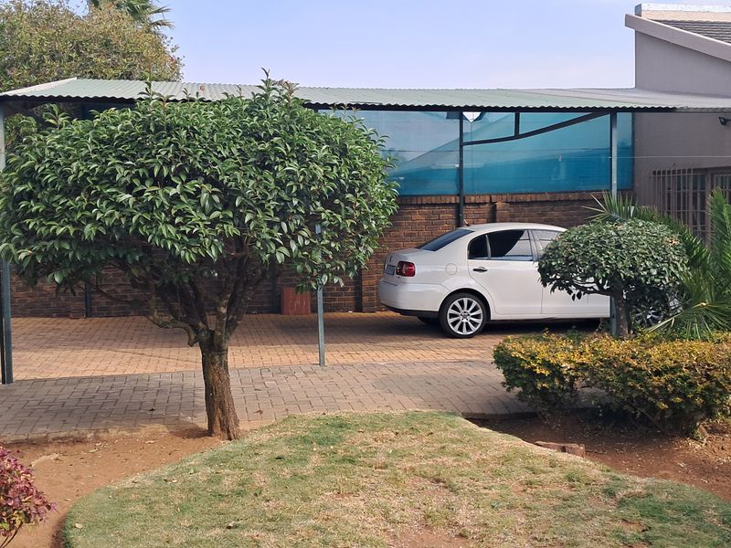 A rear exterior view of the cottage featuring a paved driveway, a covered carport structure, and established garden landscaping.